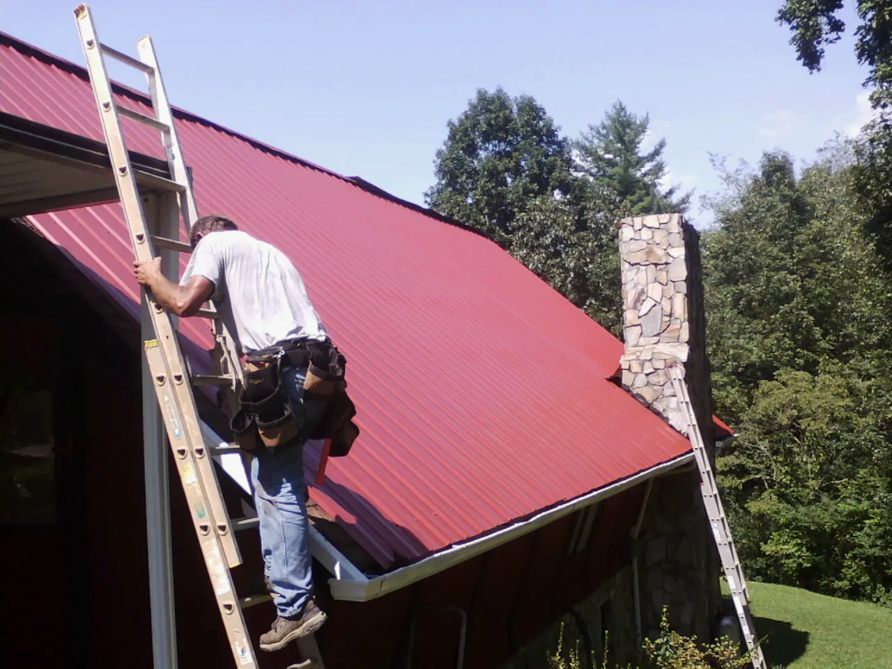 Roofer on a ladder beside a red metal roof and stone chimney — Soffit & Fascia Repair by Roofing Brothers in Gypsum