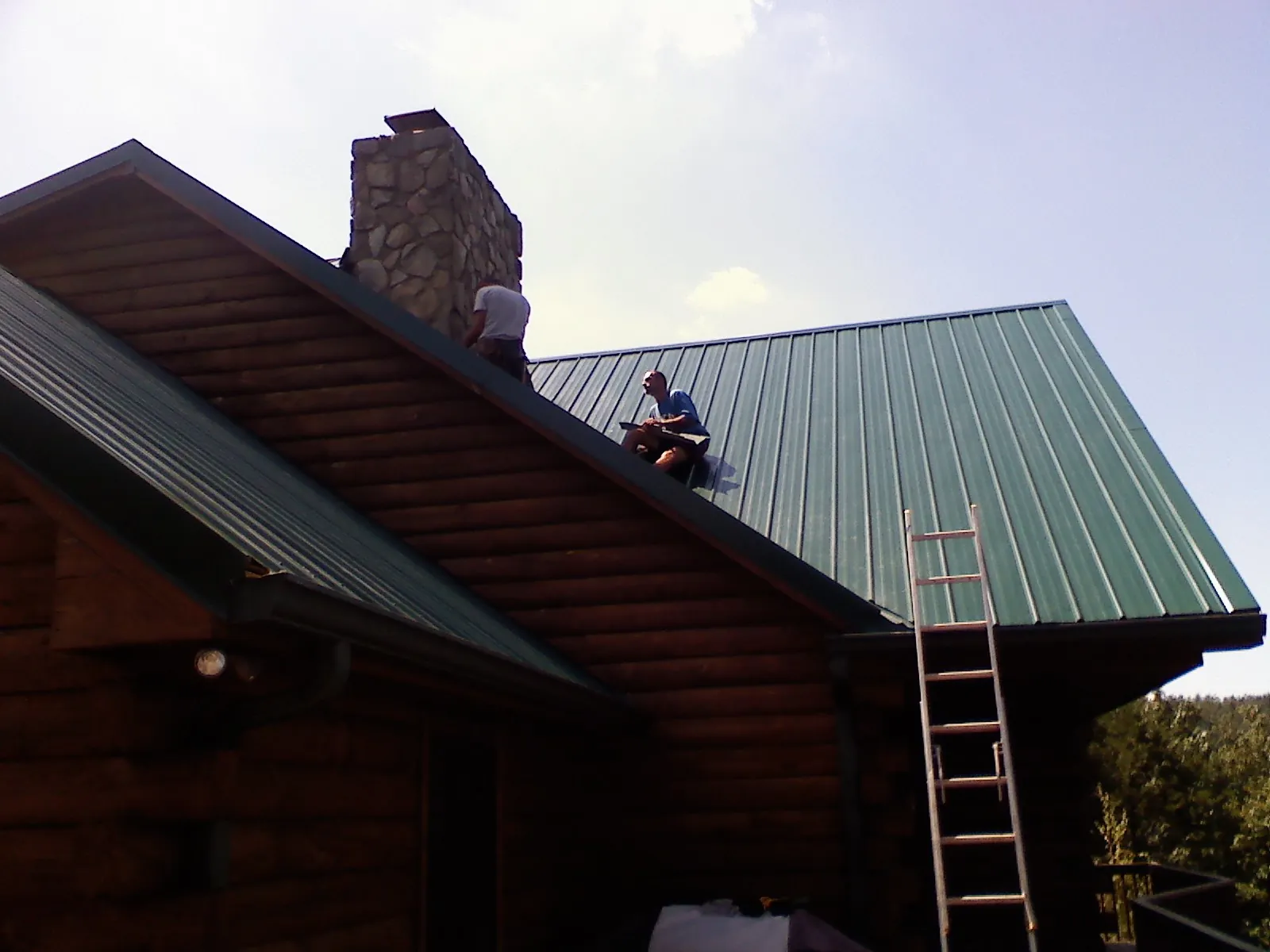 Roofers installing green metal panels beside a stone chimney on a log home in Gypsum